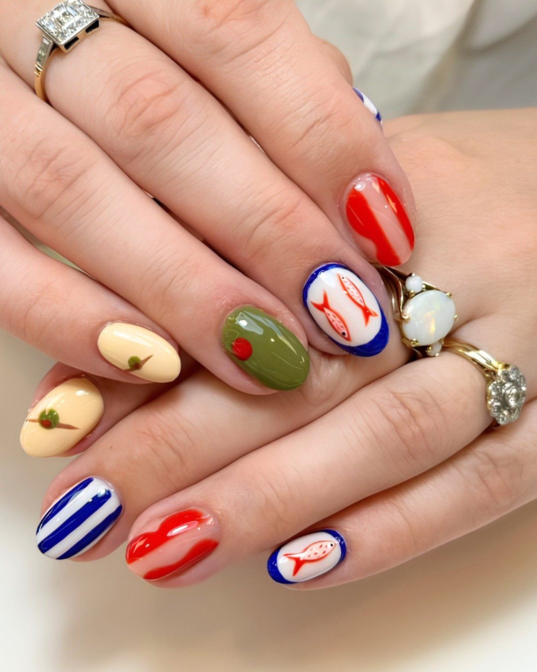 Manicured nails with red, white, and blue nautical patterns and gold rings.
