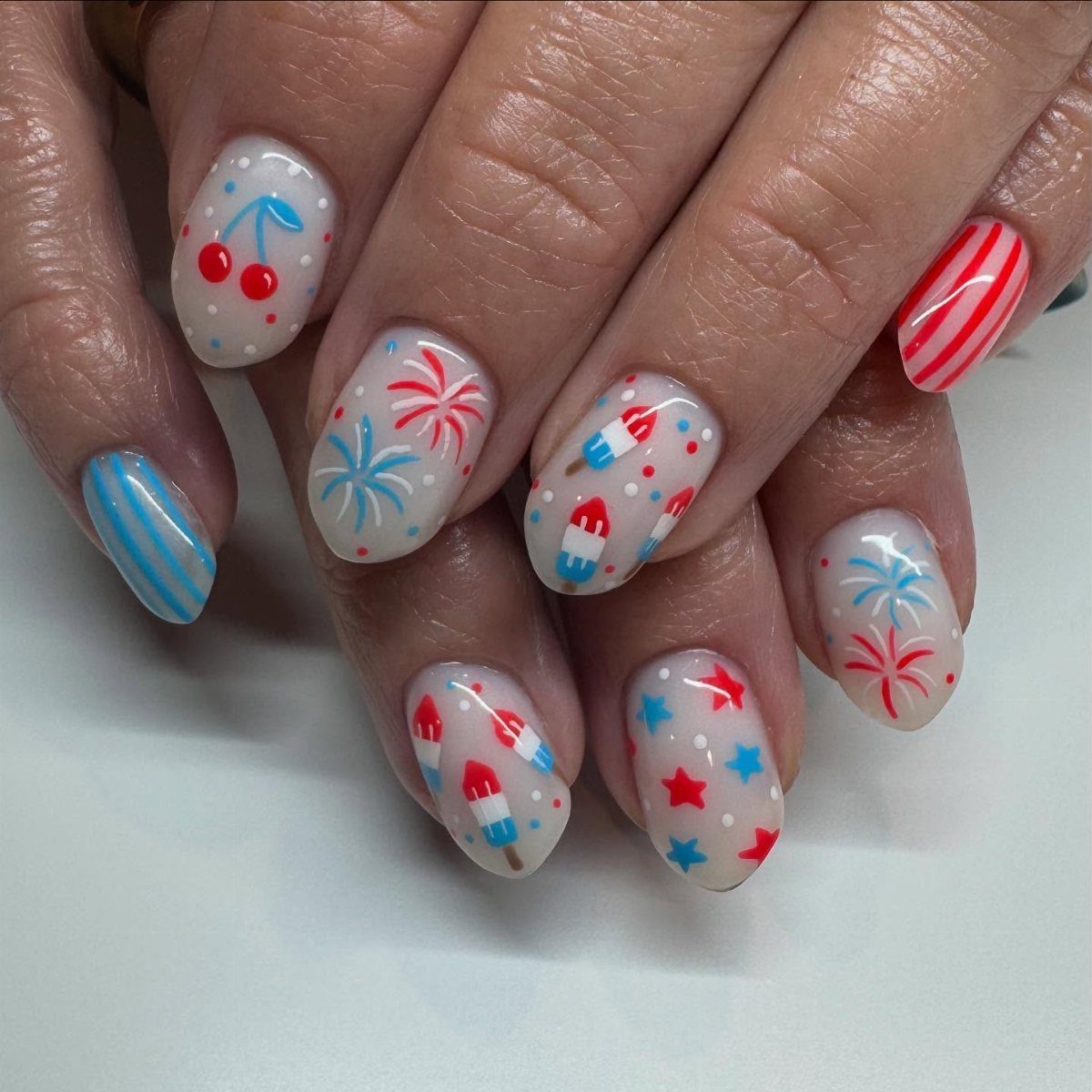 Patriotic nail art featuring fireworks, cherries, stripes, and popsicles in red, white, and blue.