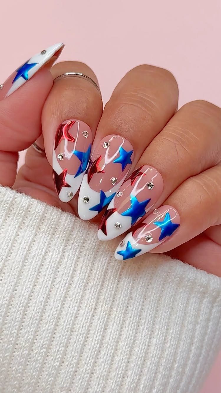 Almond nails with white tips, metallic red and blue stars, and silver rhinestones.