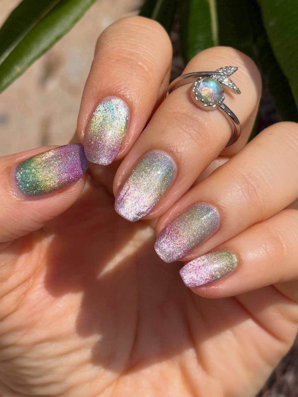 Holographic gel nails shimmering in rainbow colors on a hand wearing a ring.