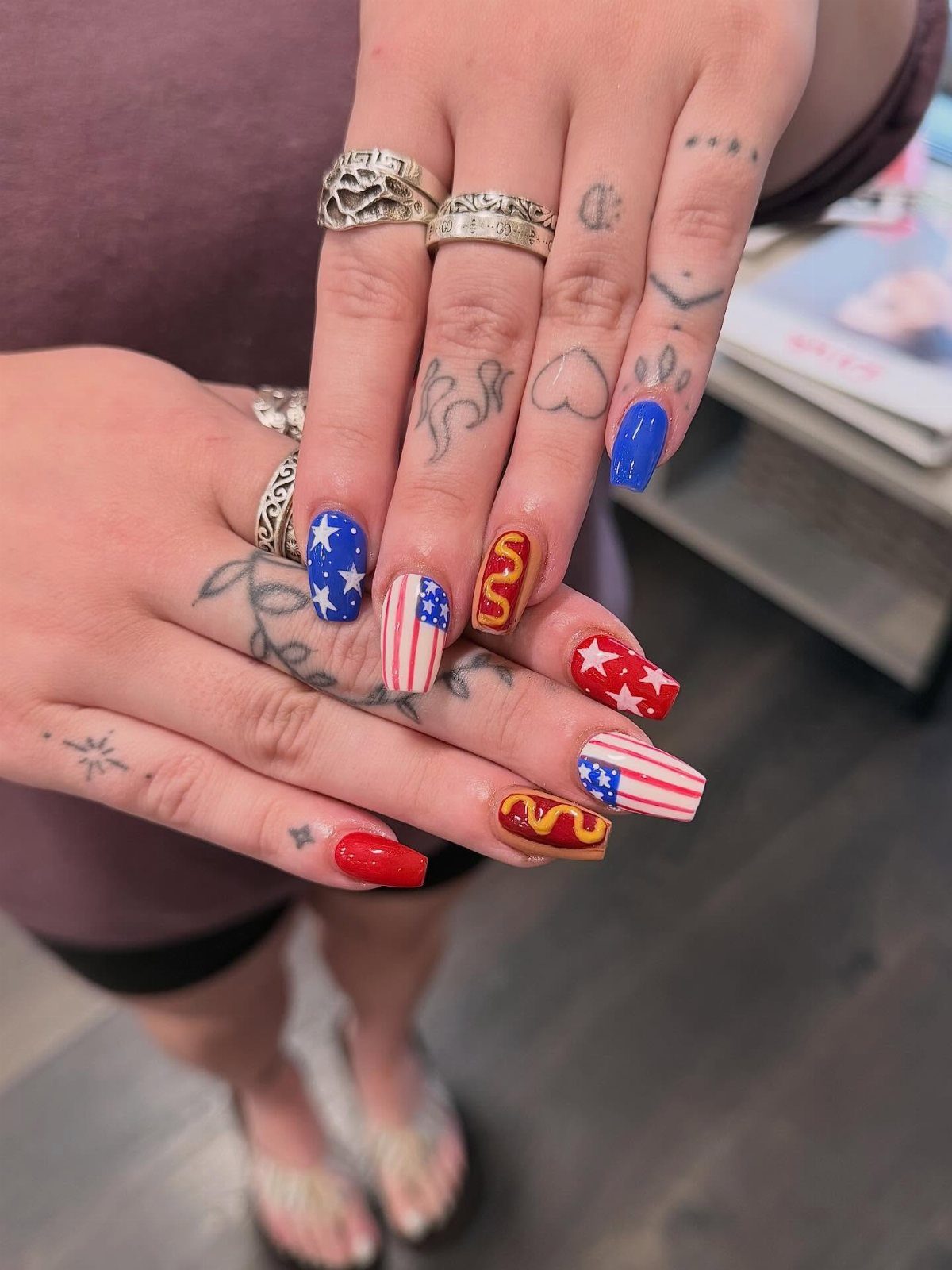 Patriotic gel nails with stars, stripes, and hot dog art on tattooed hands.