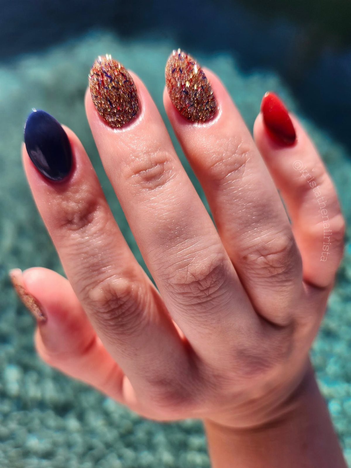 Hand with red, gold glitter, and dark blue almond-shaped gel nails.