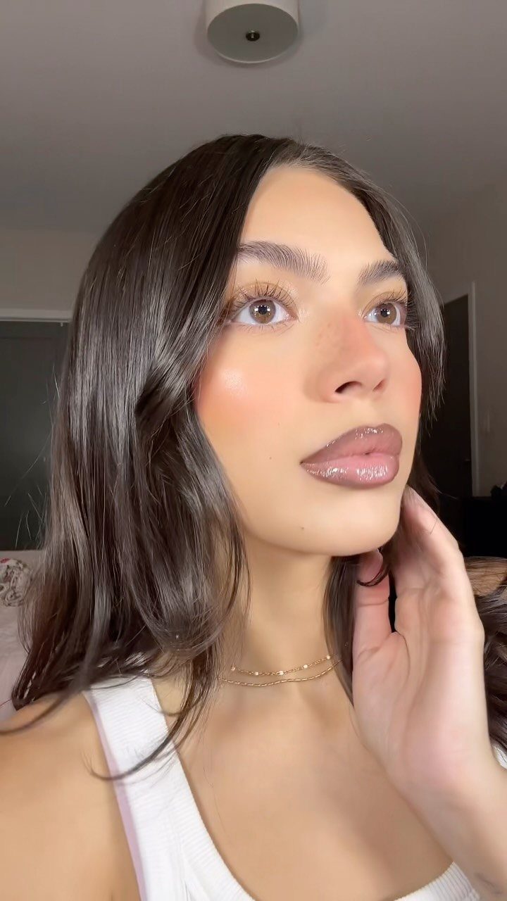 Woman with glowing skin, neutral eyeshadow, and glossy nude-pink lips posing indoors.
