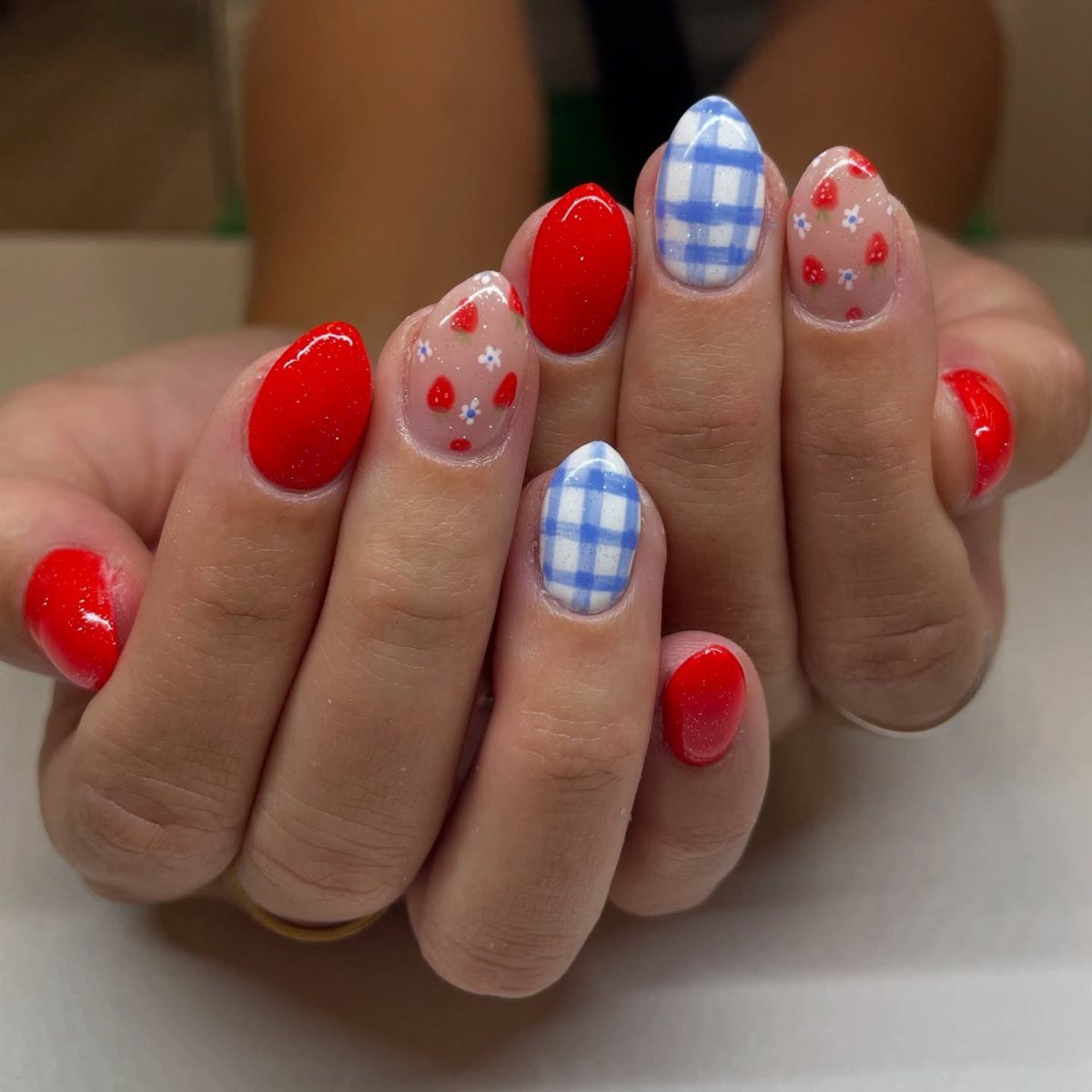 Manicured nails featuring red glitter, blue and white gingham, and strawberry patterns.