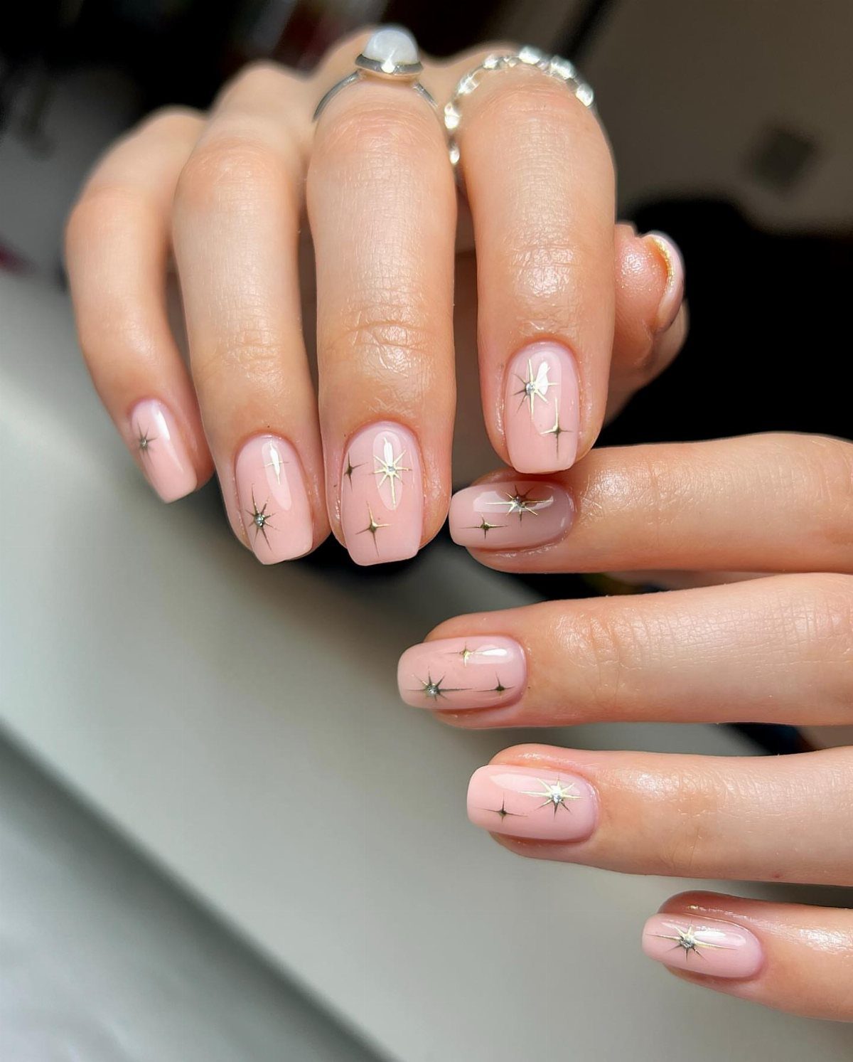 Light pink manicured nails with silver starburst designs and silver rings on fingers.