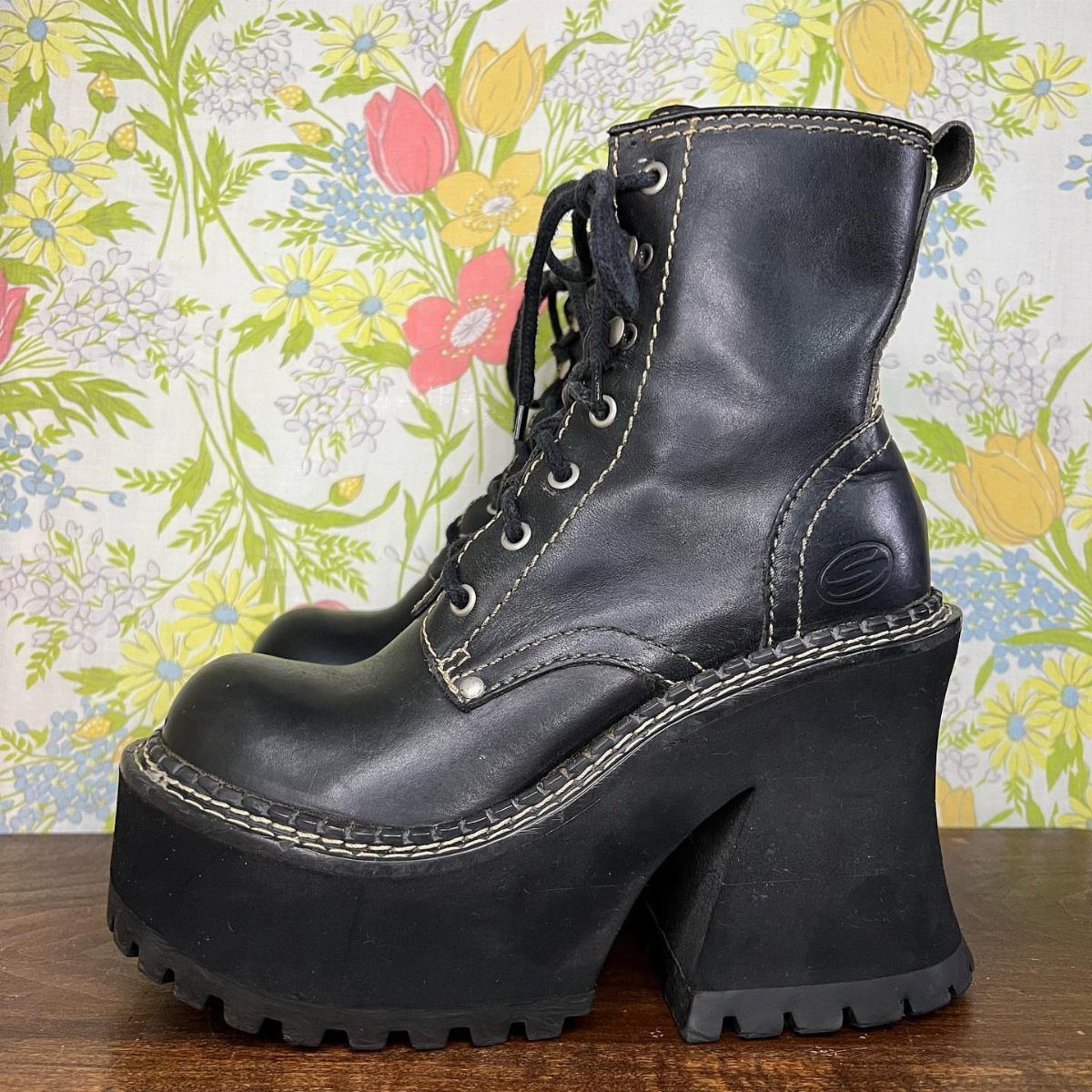 Black leather lace-up boots with massive chunky platform soles against floral wallpaper.