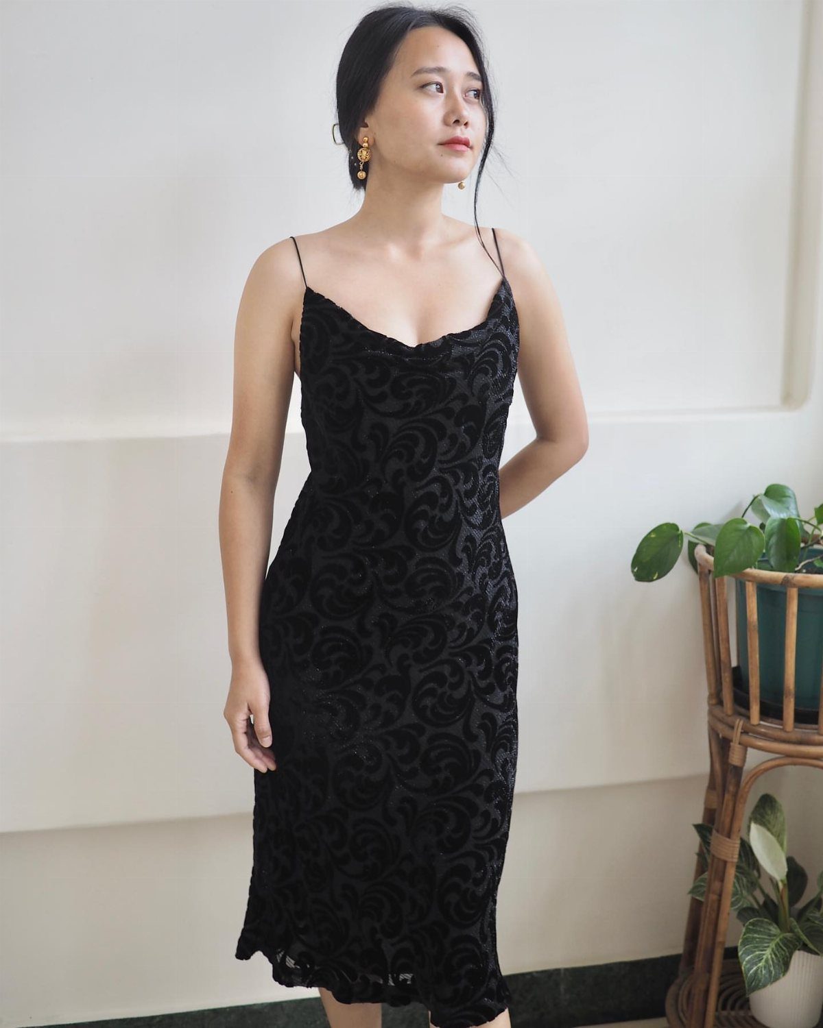 Woman wearing a black velvet midi dress with thin straps against a white wall.