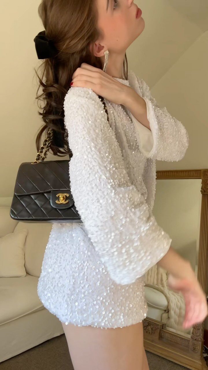 Woman in a white sequined dress and blazer with a black quilted handbag.