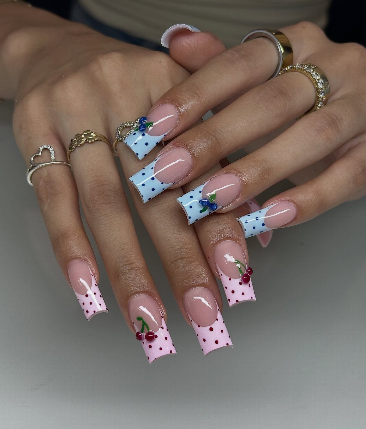 Long acrylic nails with blue and pink polka dot tips and fruit charms.