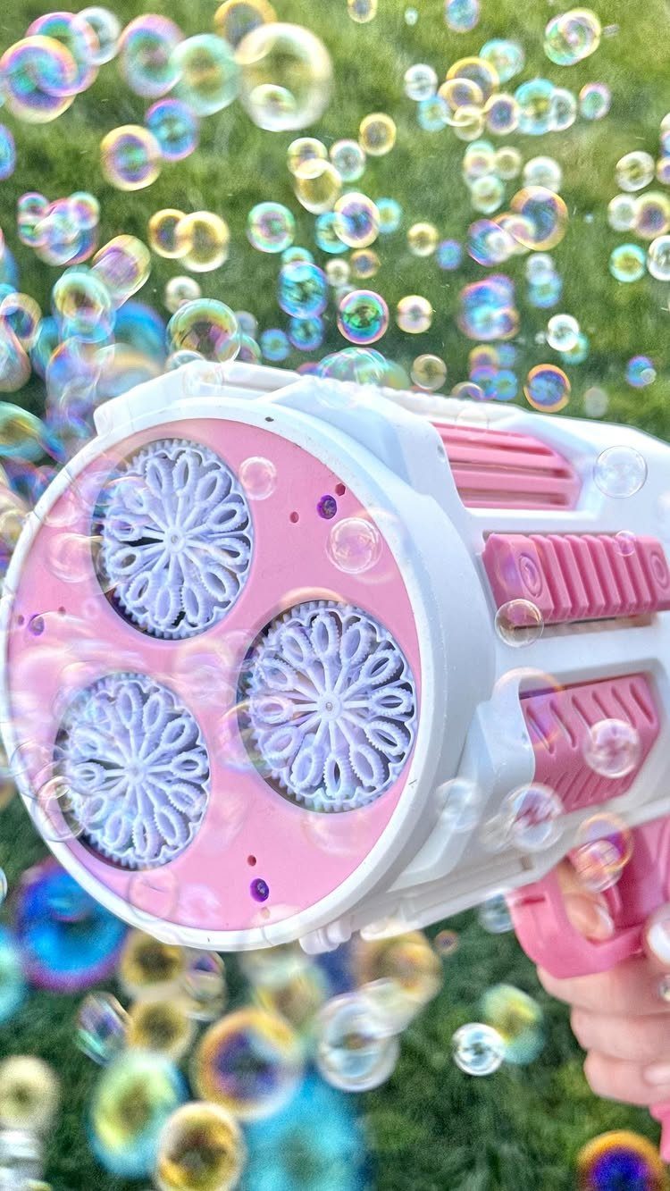 Pink and white bubble blower machine creating many iridescent bubbles over green grass.