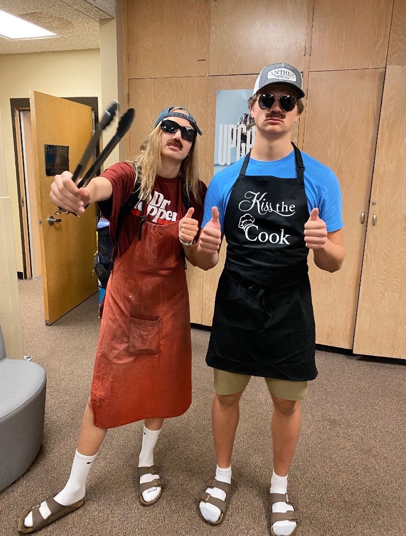 Two people wearing aprons over t-shirts and sunglasses posing as BBQ dads.