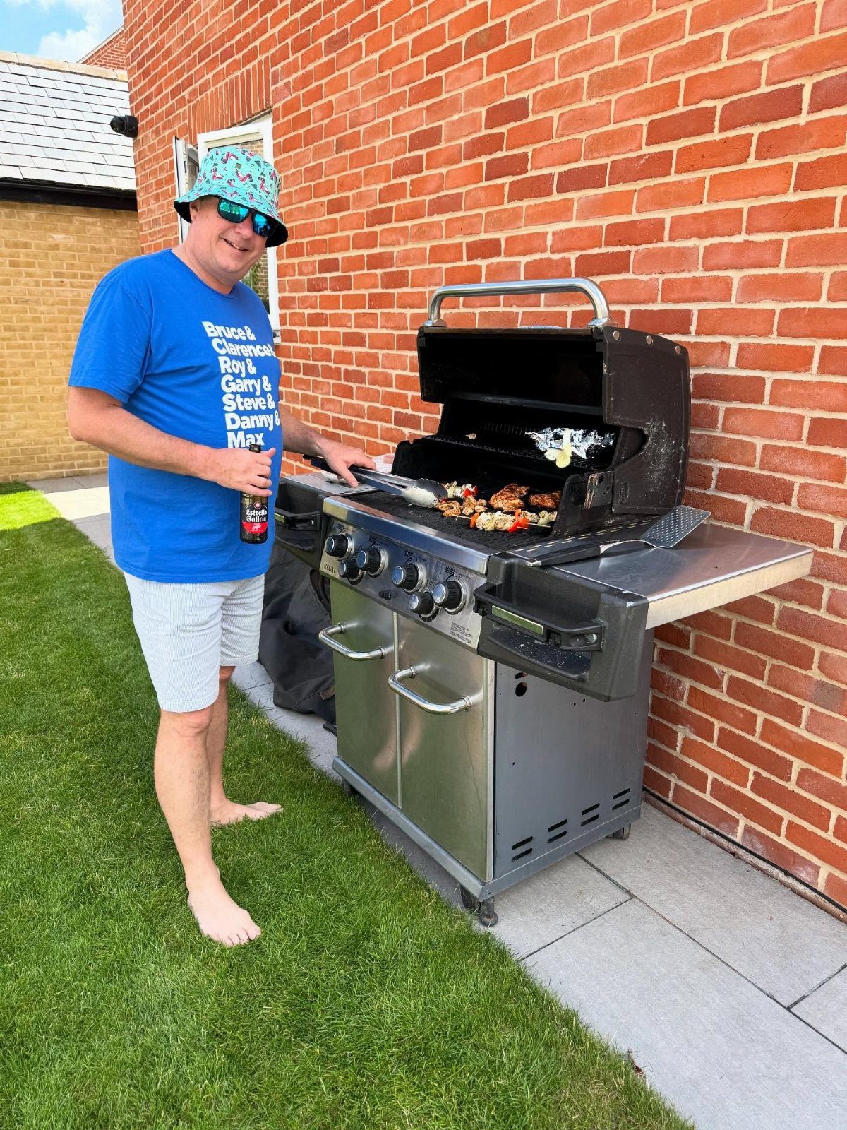 Man in a patterned bucket hat grilling food on a gas barbecue grill.