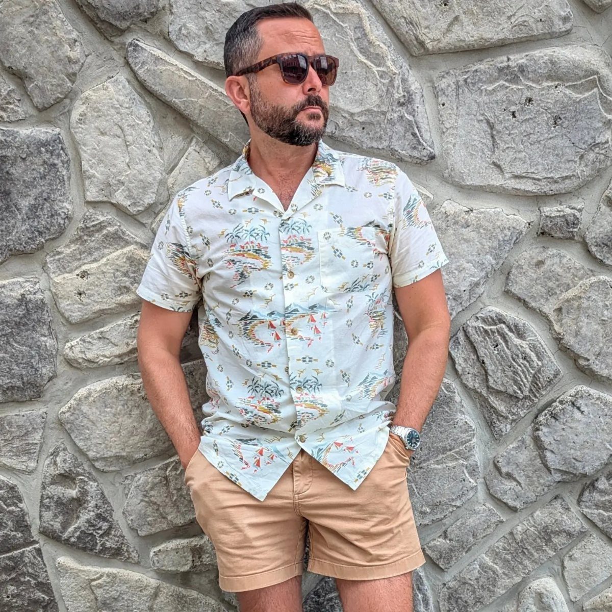 Man wearing a white tropical print Hawaiian shirt and tan shorts by stone wall