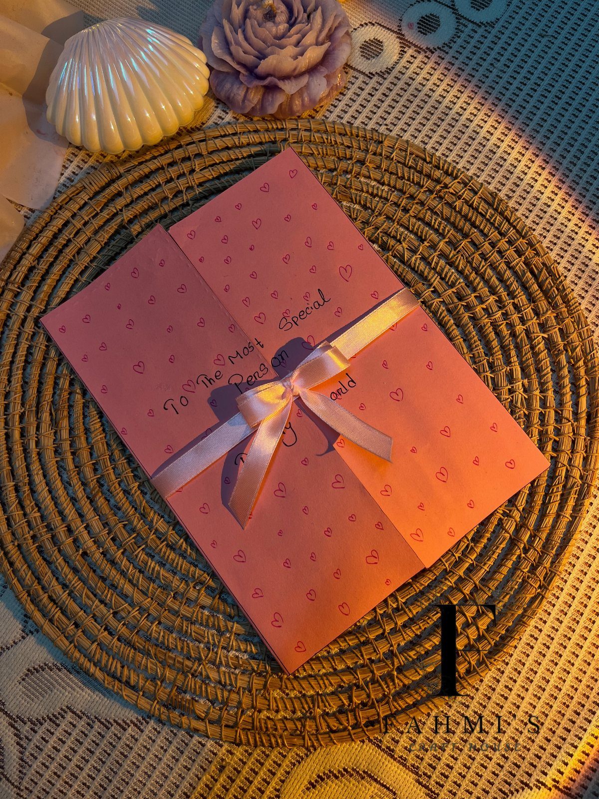 Pink heart-patterned envelope tied with a ribbon on a woven straw mat.