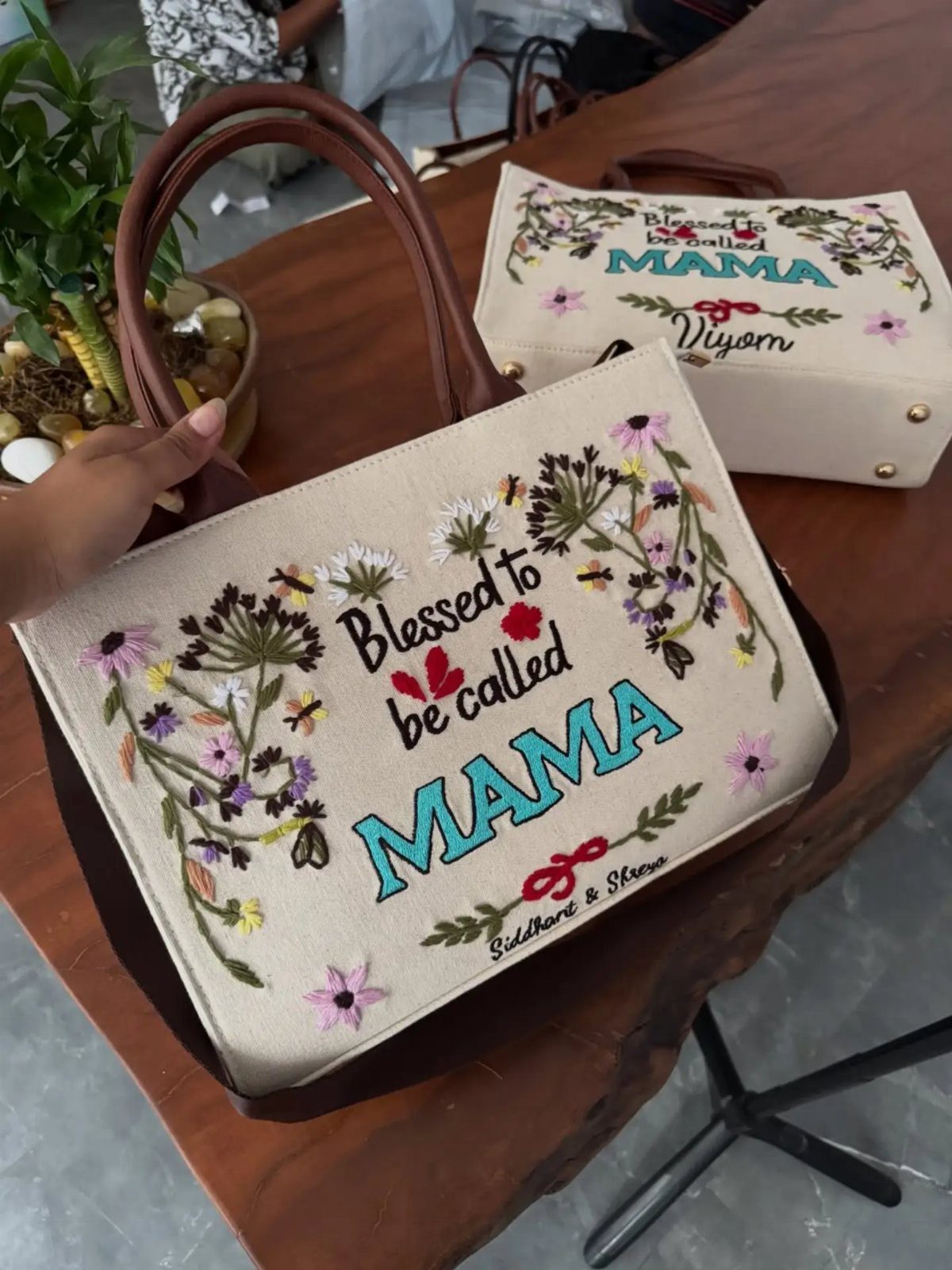 Two cream canvas tote bags with floral embroidery and 'MAMA' text on wood.