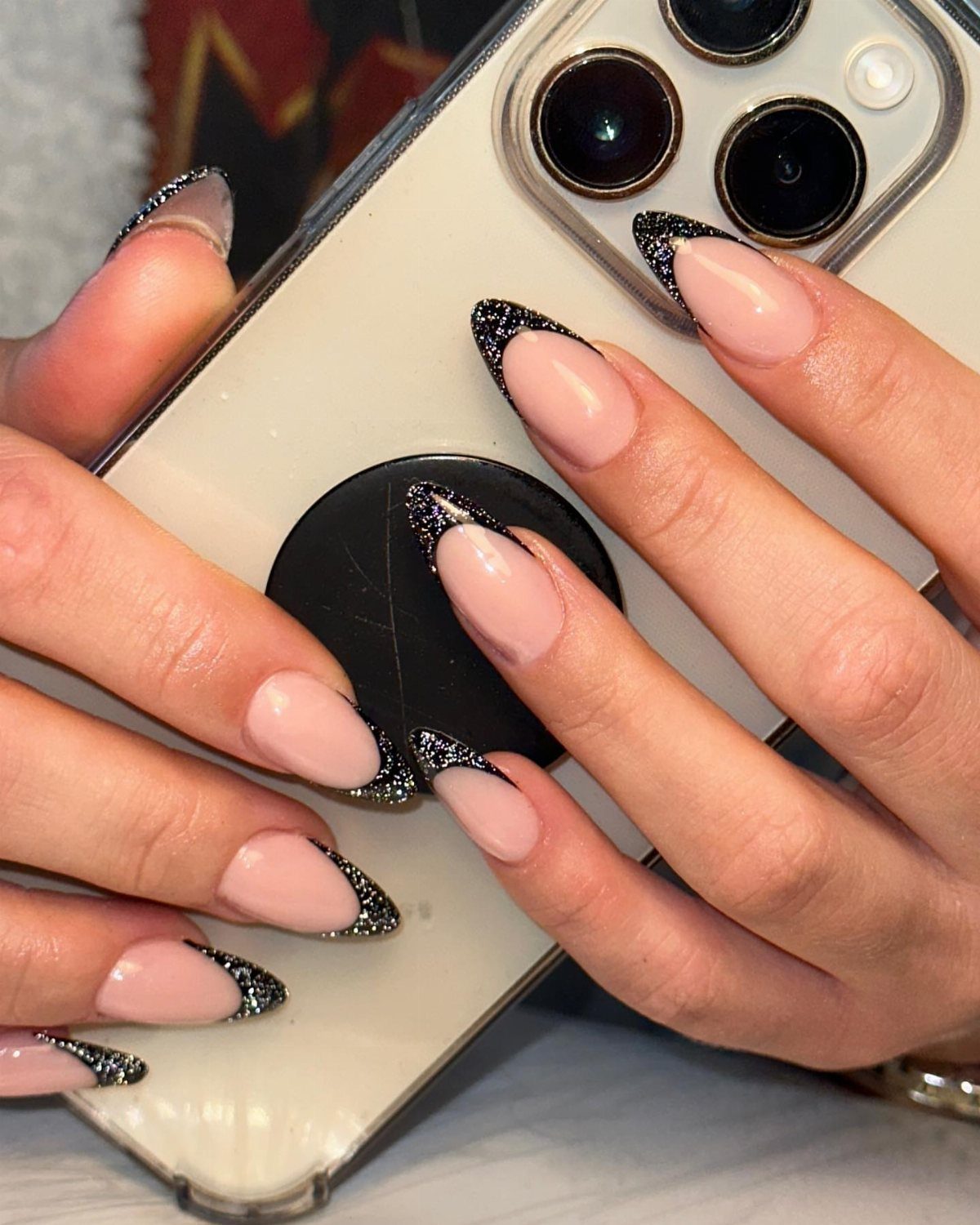 Nude almond nails with black glitter French tips holding a white smartphone