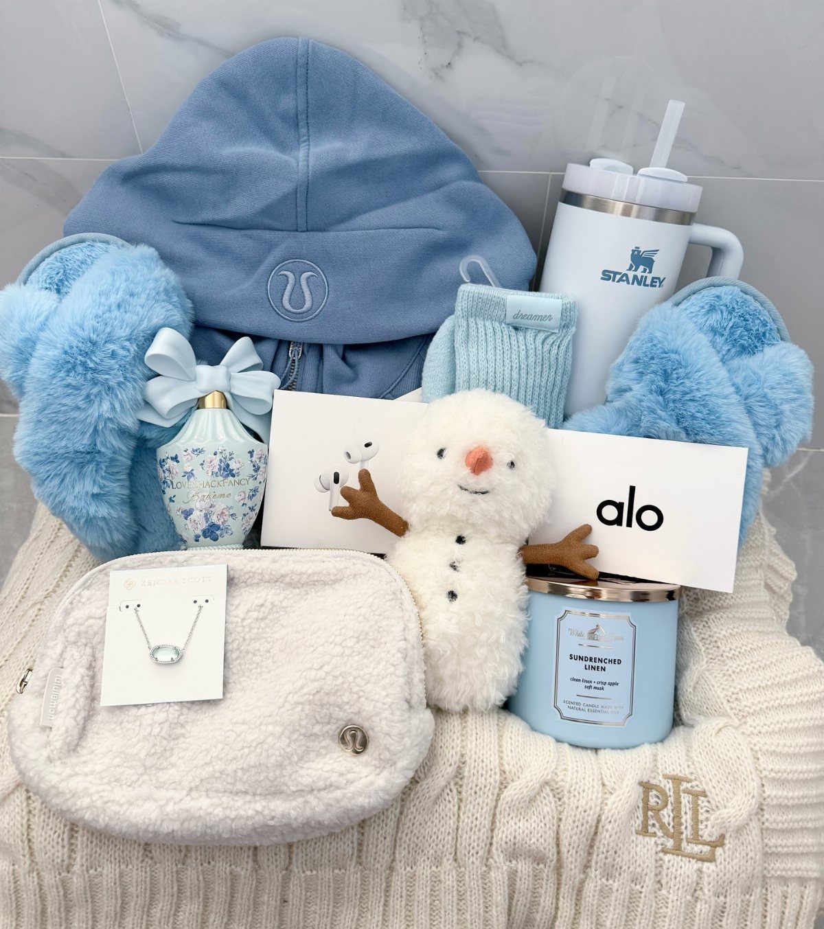 Gift set with blue accessories, a plush snowman, and a white cable-knit sweater.