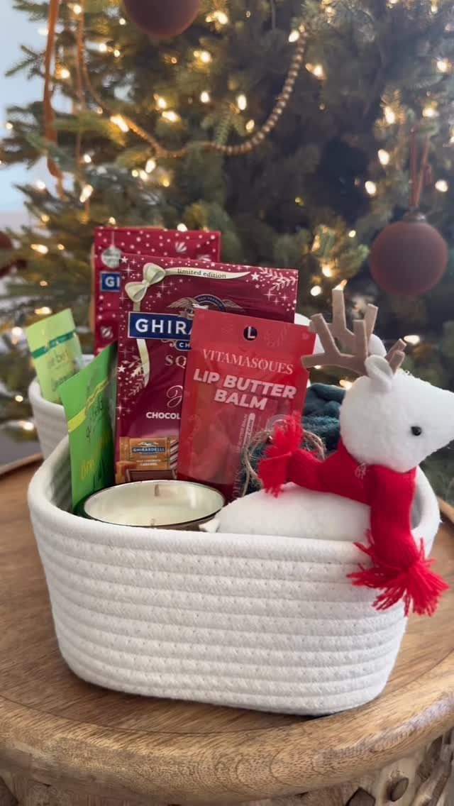White woven gift basket with chocolates, lip balm, and a plush reindeer near Christmas tree.