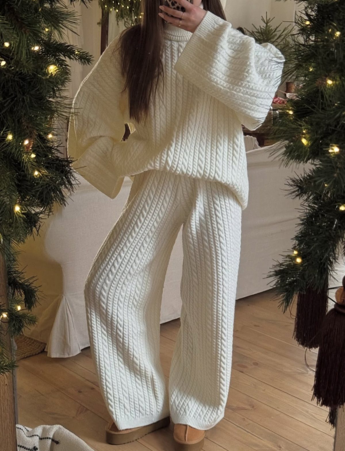 Person wearing a cream cable-knit lounge set standing between lit Christmas trees.