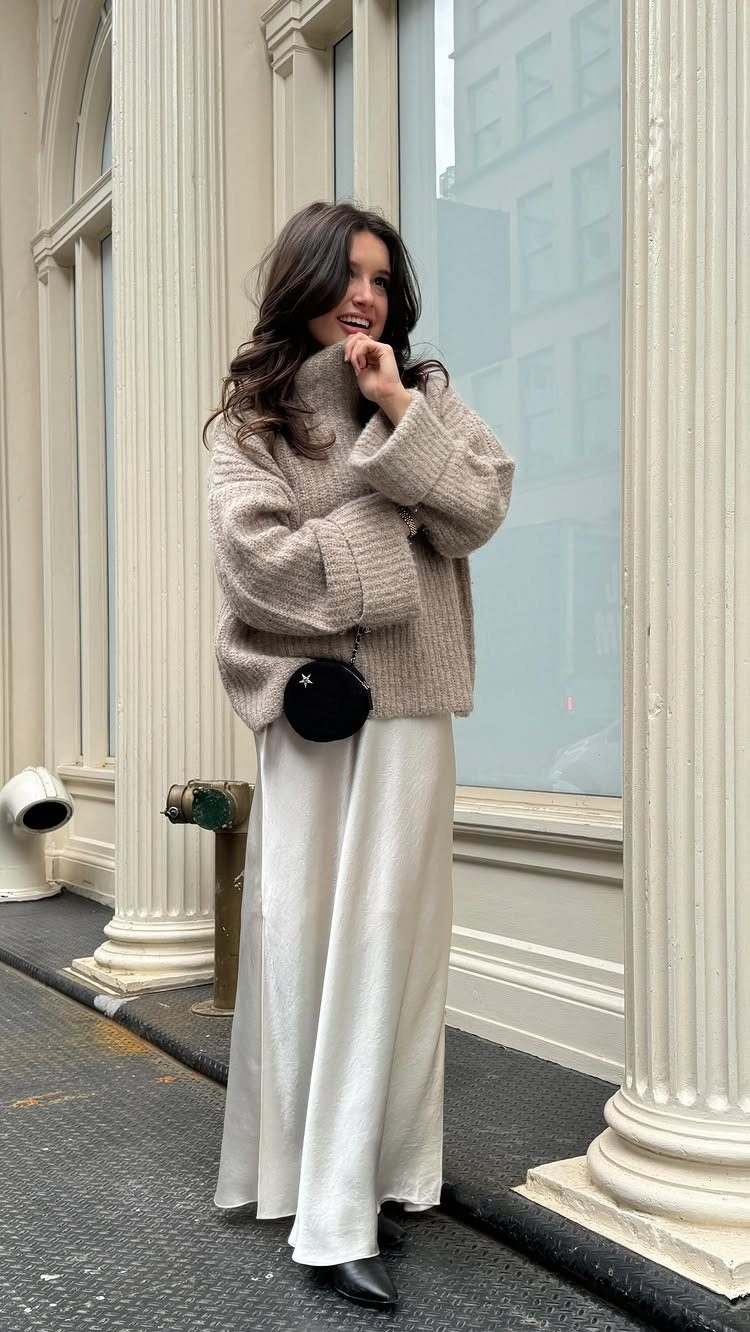Woman wearing a beige chunky knit sweater and a long white satin skirt.