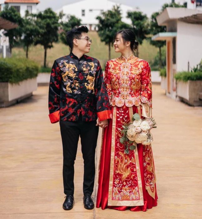 Couple in traditional red and gold Chinese wedding attire holding a white bouquet