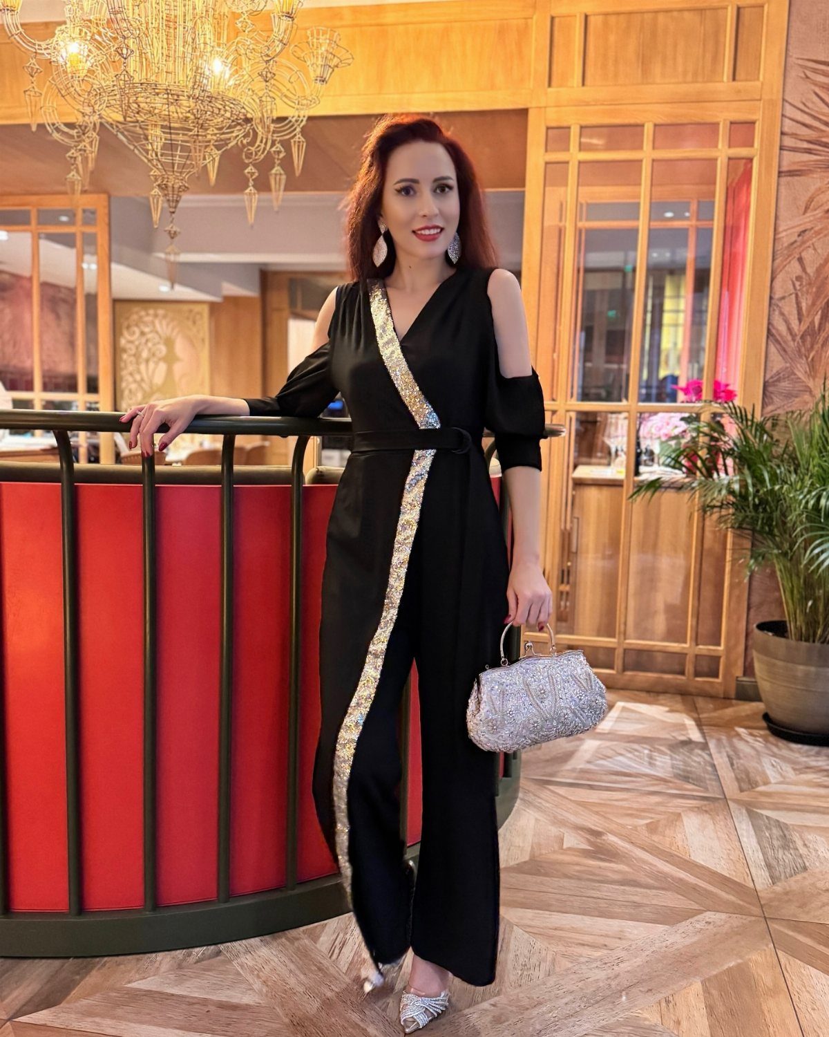 Woman wearing a black jumpsuit with gold trim and silver glittery handbag.