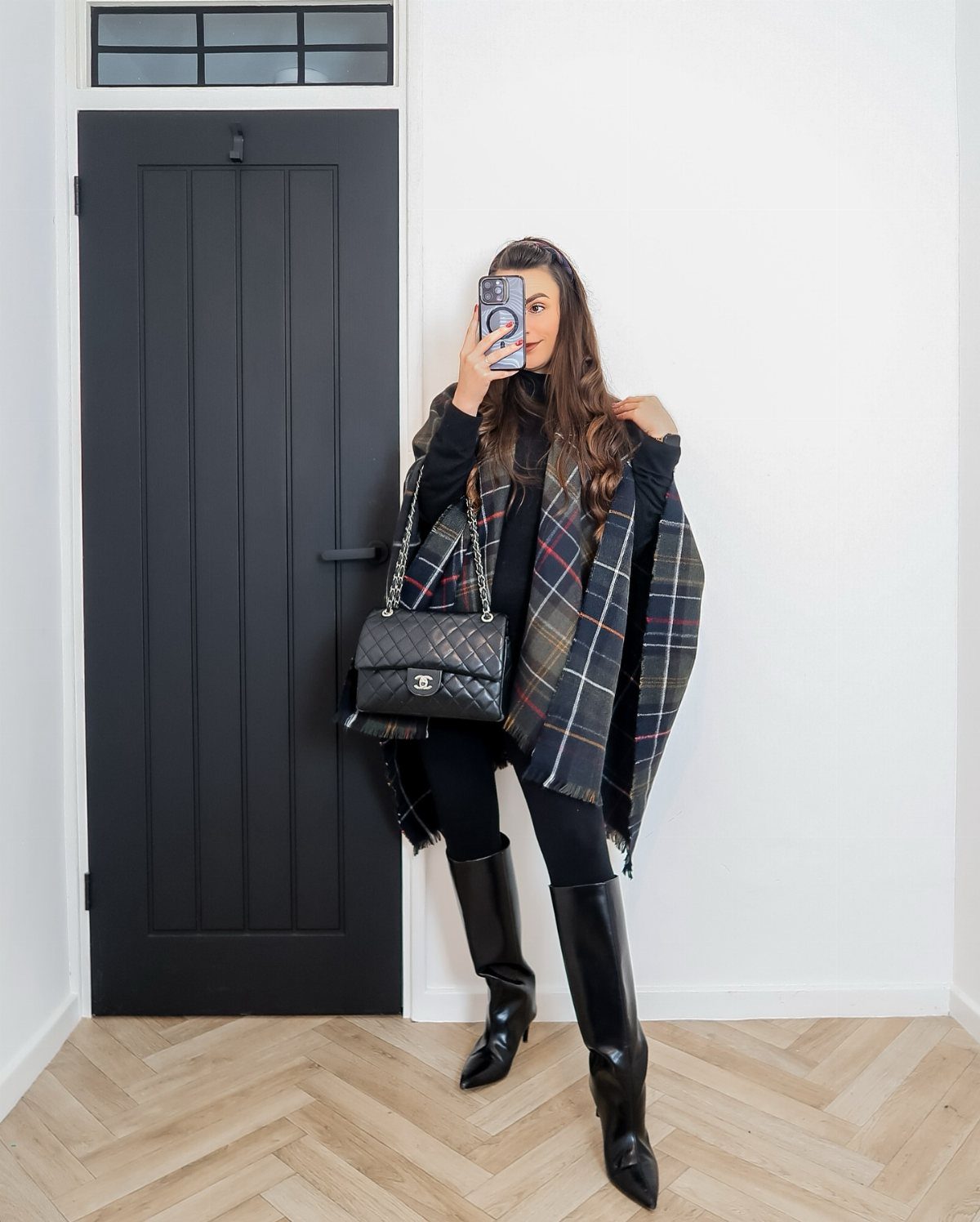 Woman wearing black leather knee-high boots and a plaid coat taking a selfie.