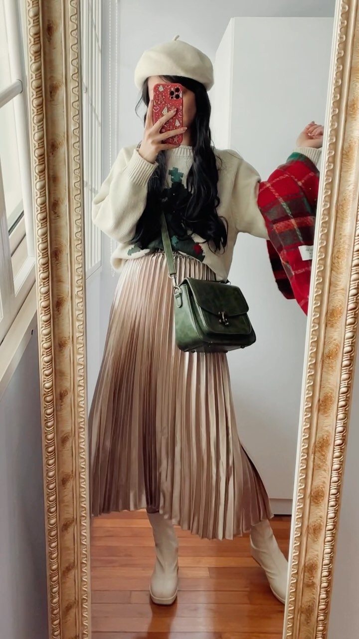 Woman in a cream knit sweater and gold pleated midi skirt taking a selfie.