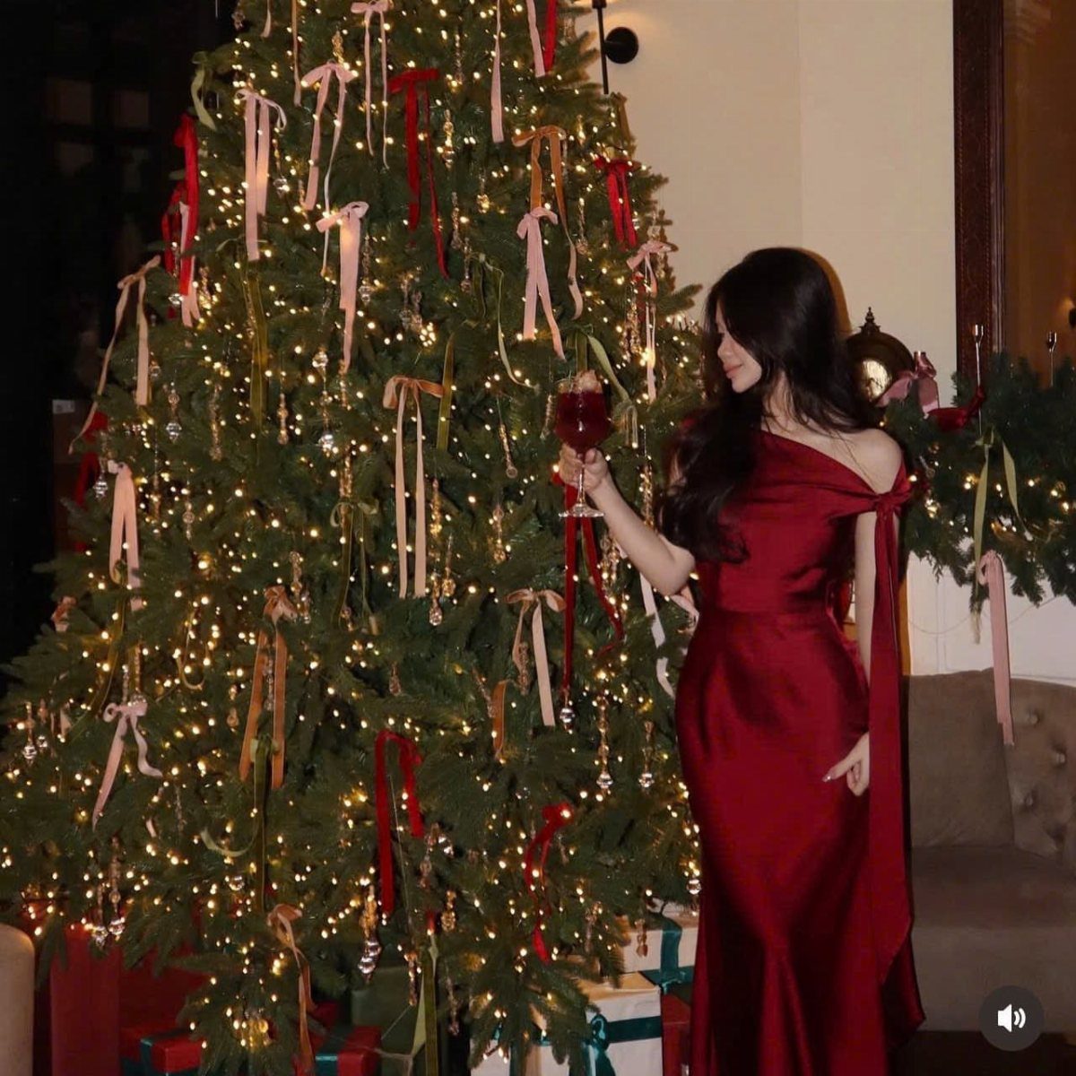 Woman in a long burgundy satin dress holding wine by a Christmas tree
