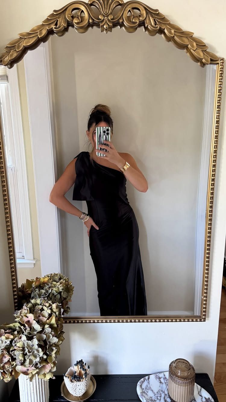 Woman in a long black one-shoulder satin dress posing in a gold mirror.