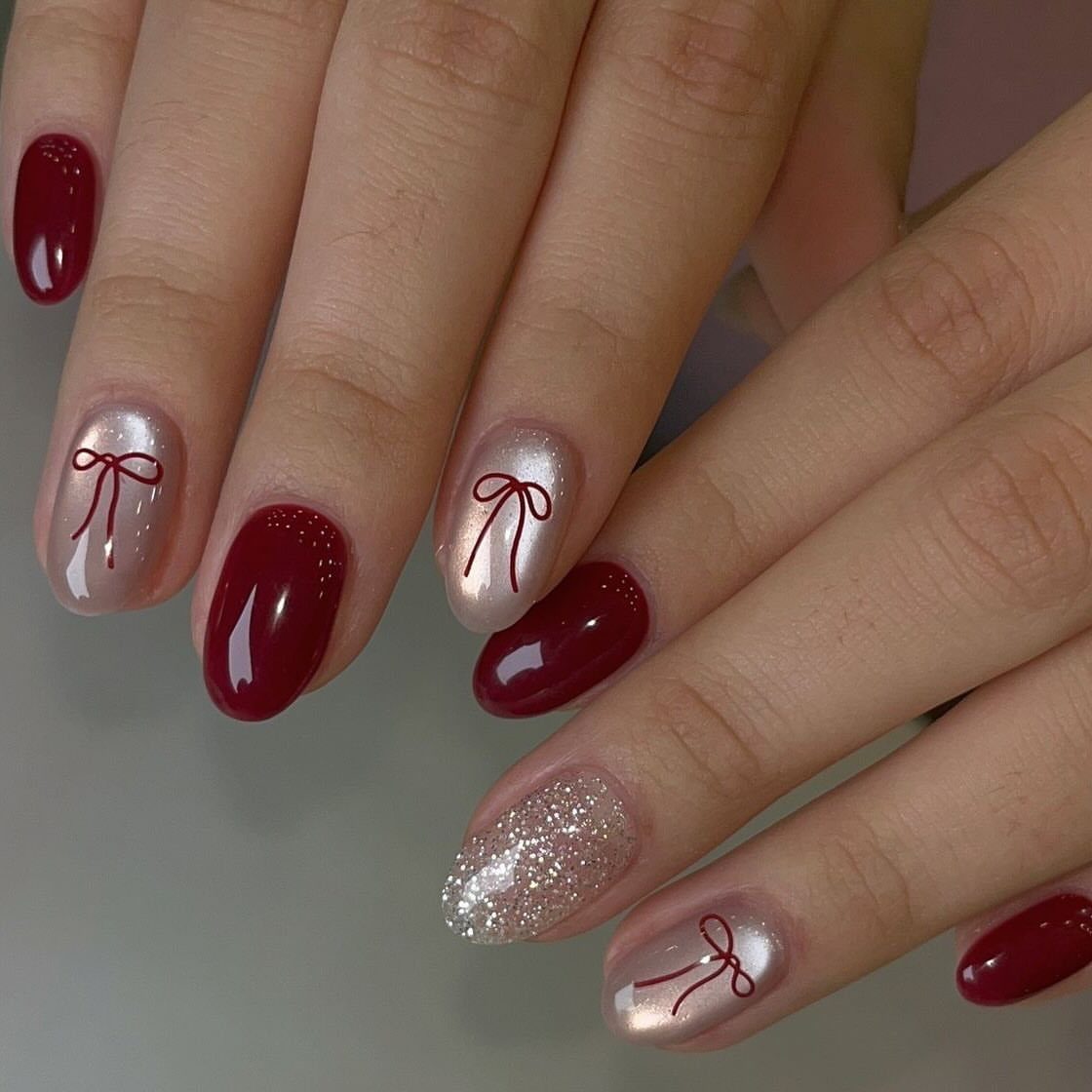 Short oval nails with deep red, champagne gold bows, and silver glitter polish.