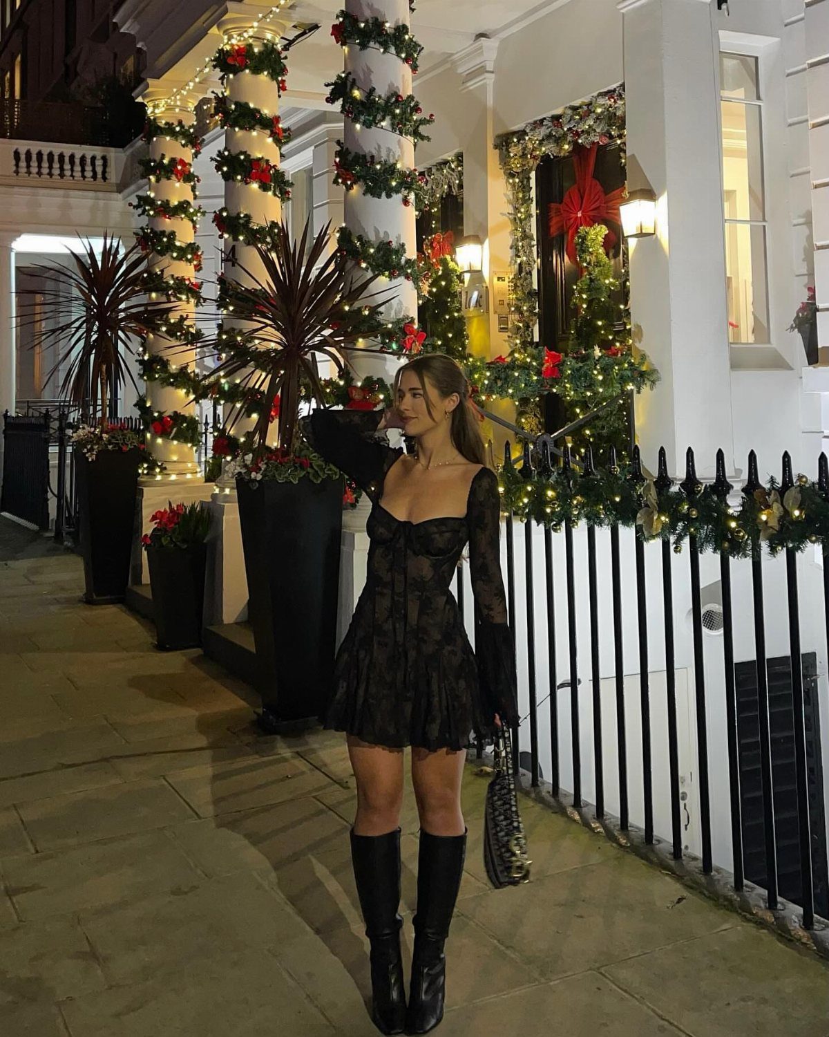 Woman in black mini dress and knee-high boots standing by Christmas decorations.