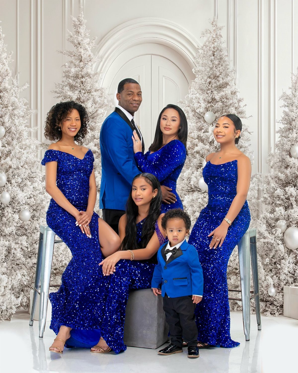 Family in royal blue formal outfits posing between white Christmas trees in a studio.