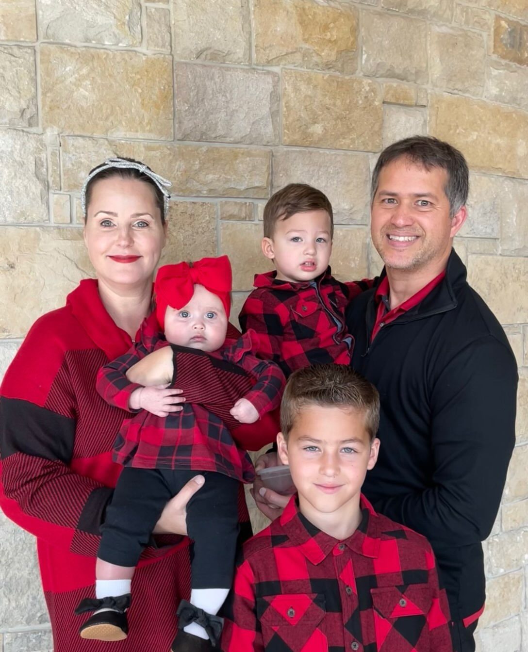 Family posing in red and black buffalo plaid outfits against a stone wall.