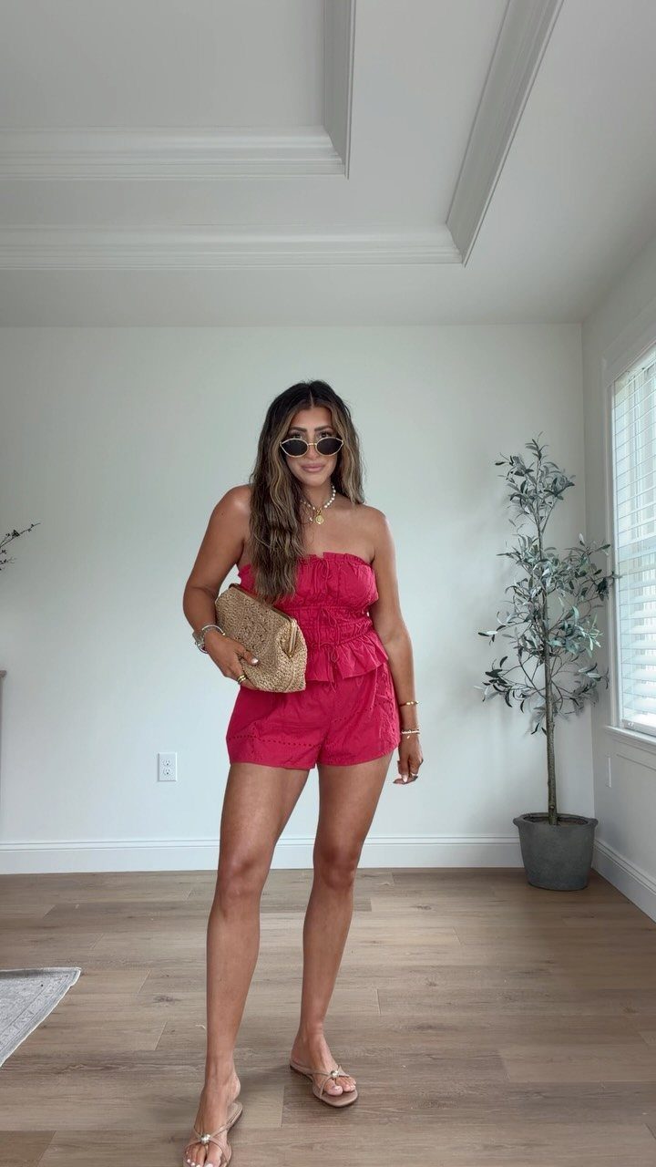 Woman wearing a red strapless romper and sunglasses standing in a white room.