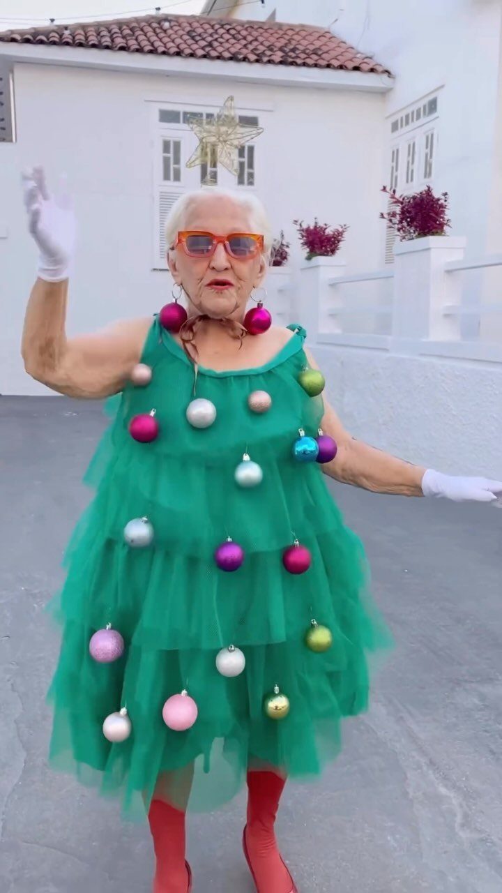 Elderly woman in a green ornament-decorated dress and red boots posing outdoors.