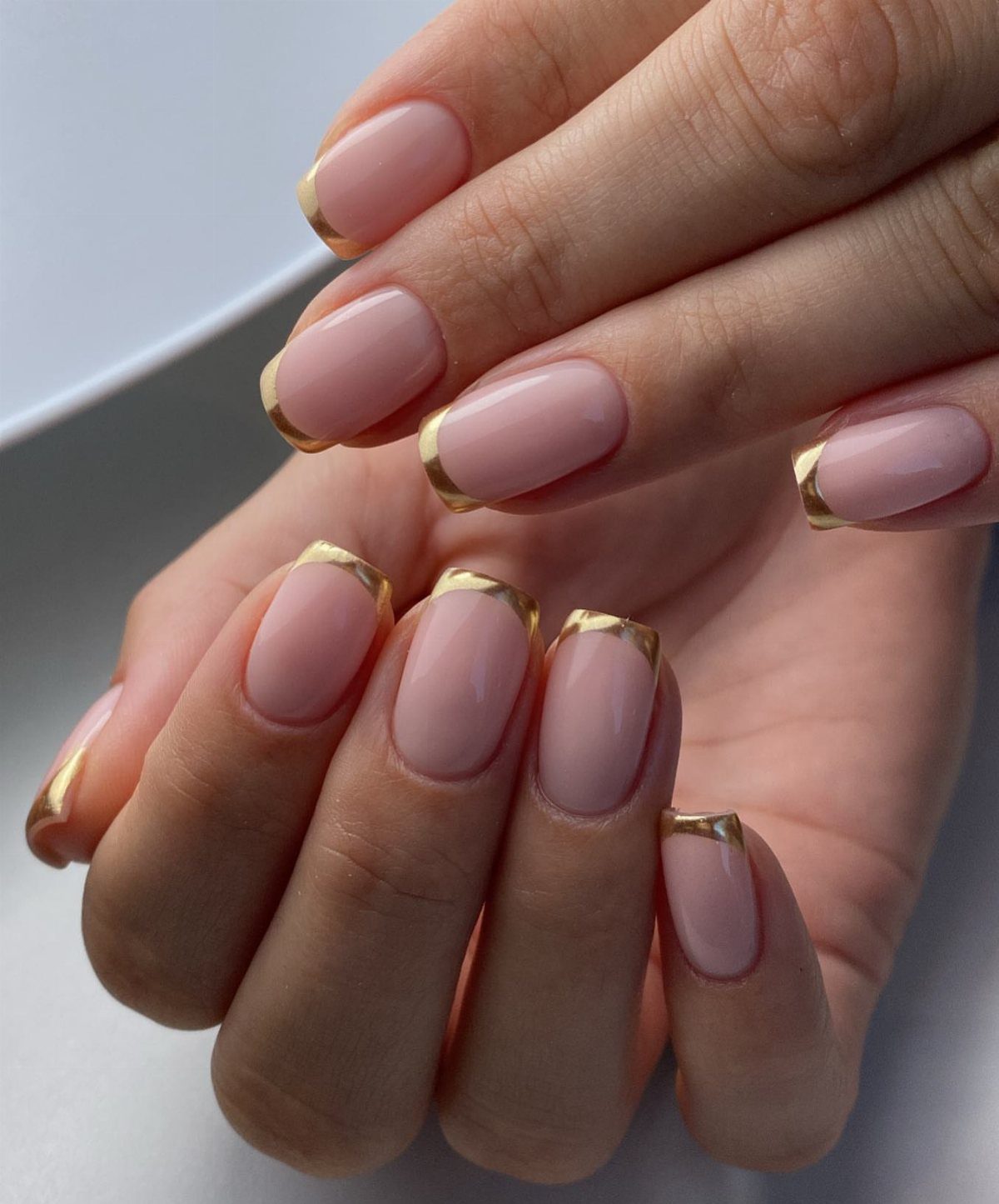 Nude pink oval nails featuring metallic gold French tips on two hands.