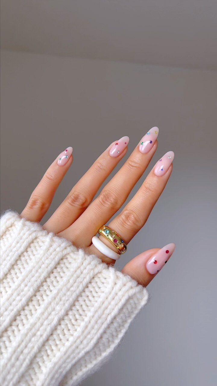 Nude almond nails decorated with small multi-colored rhinestones and a white sweater sleeve.