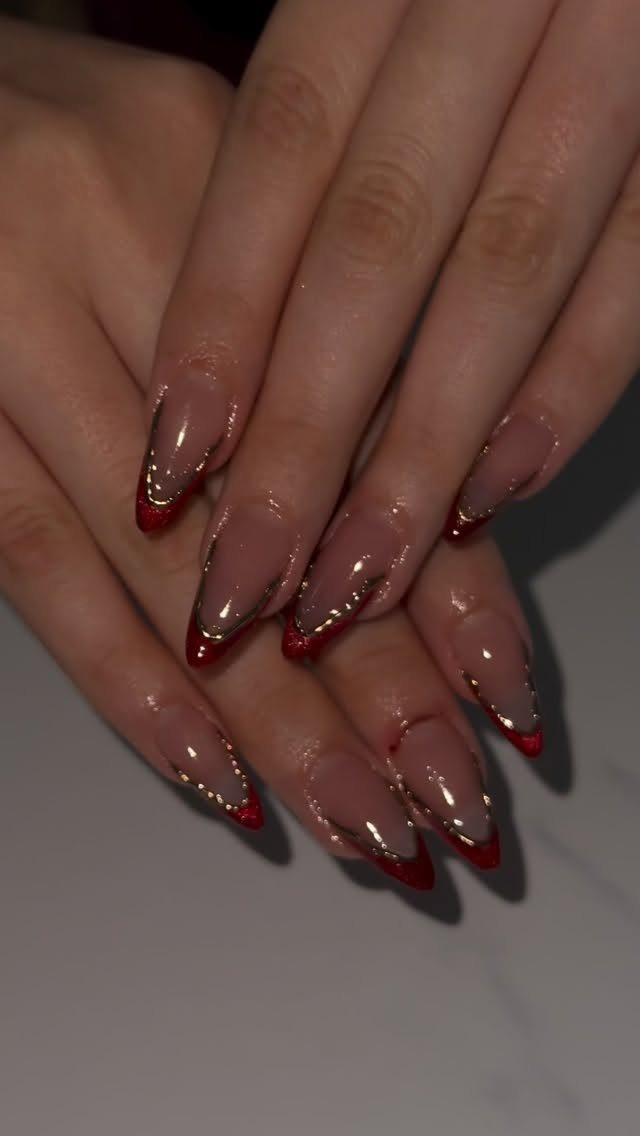 Almond-shaped nails with nude base, red tips, and gold glitter accents.