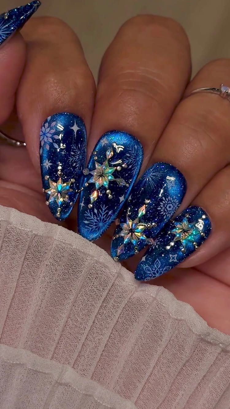 Blue shimmering nails with white snowflake patterns and iridescent crystalline snowflake charms.