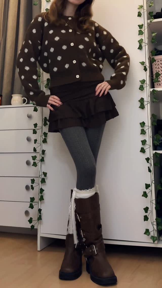 Person in brown polka dot cardigan, ruffled skirt, grey tights, and tall brown boots.