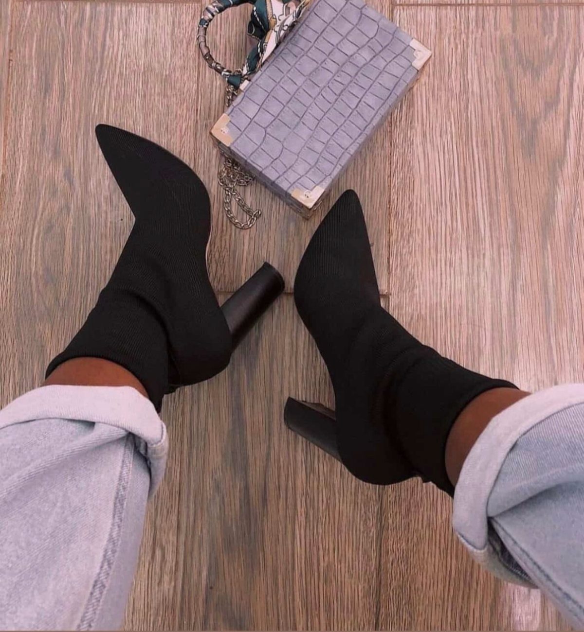 Black pointed-toe ankle boots paired with cuffed light-wash jeans on wooden flooring.
