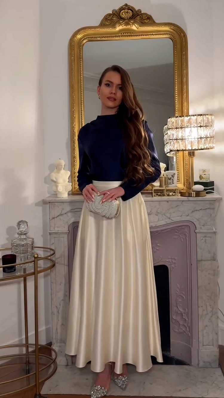Woman wearing a navy blue top and long cream satin skirt by a fireplace