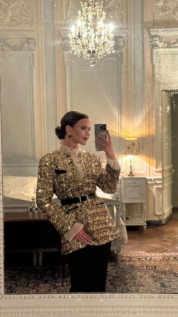 Woman in a gold sequined blazer and black pants posing in an ornate room.