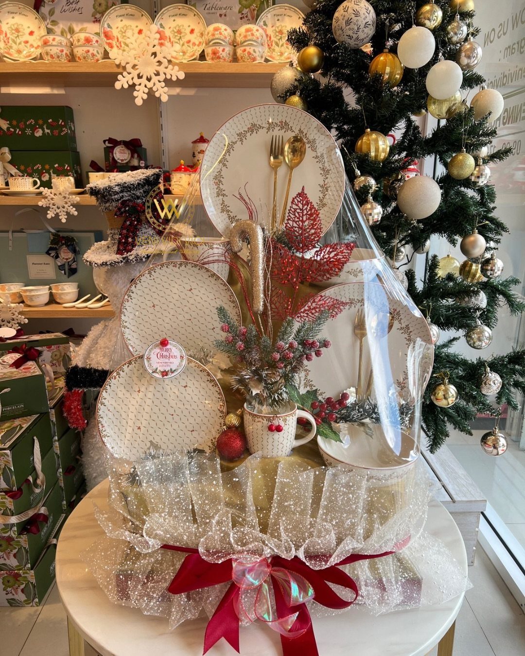 Wrapped gift basket with gold plates and cutlery next to a Christmas tree