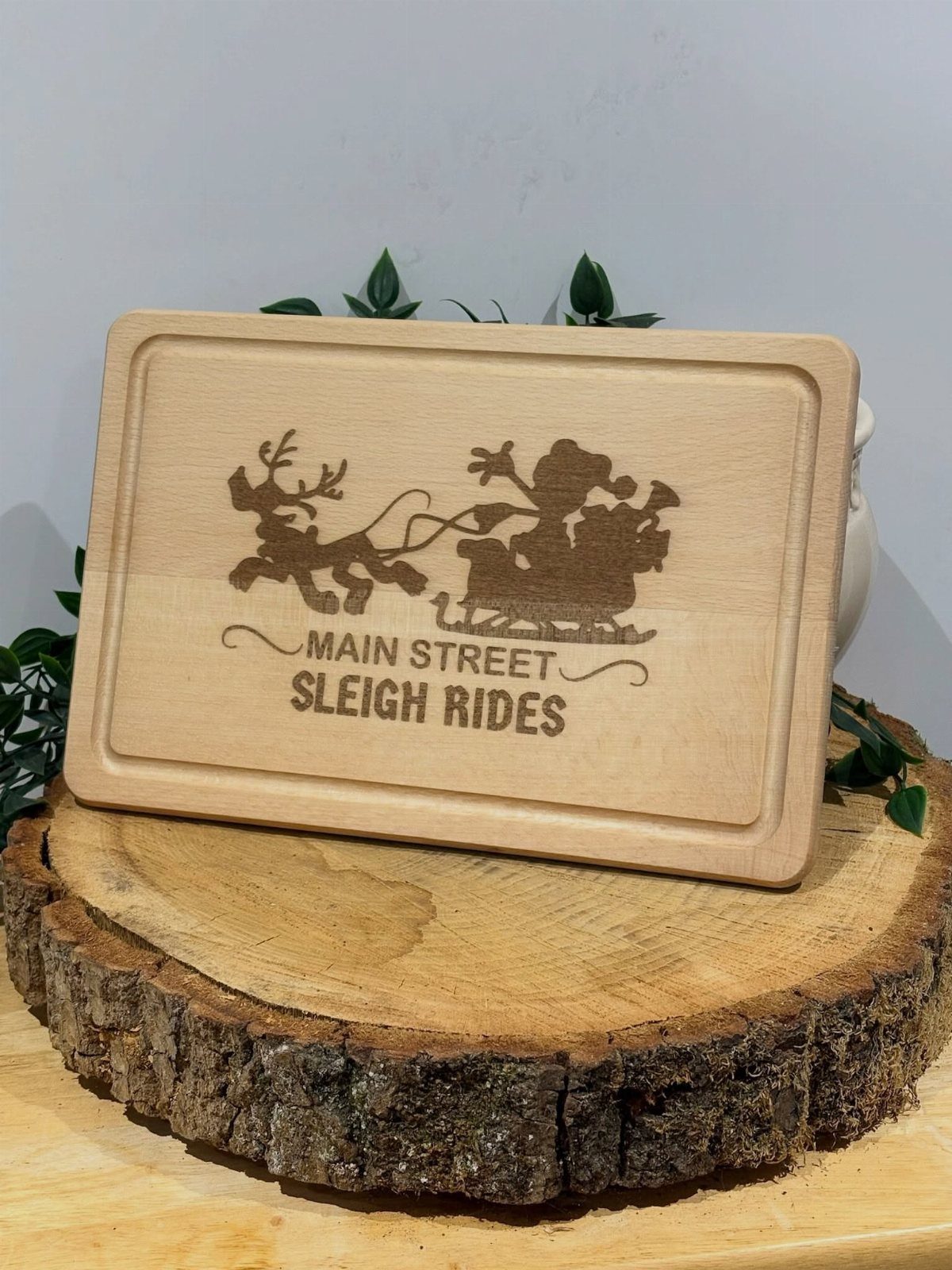 Engraved wooden cutting board with Santa's sleigh resting on a rustic log slice.