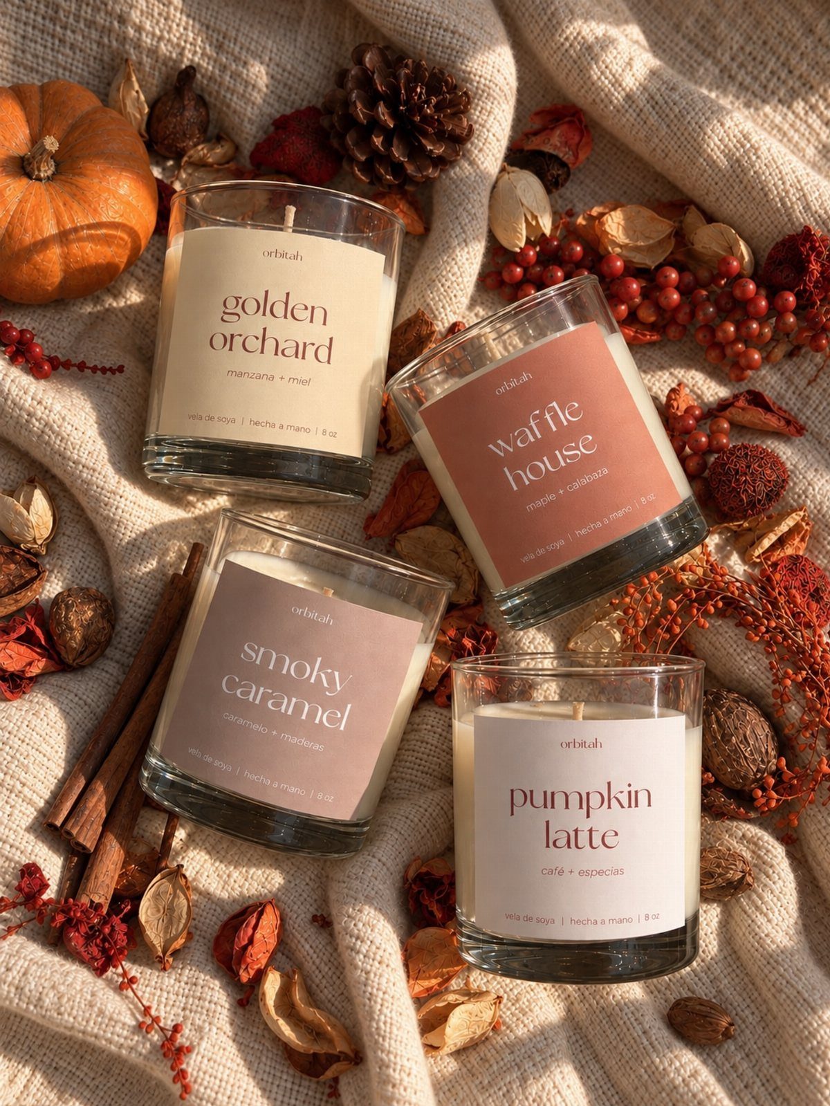 Four scented candles with autumn labels arranged on fabric with fall decorations.