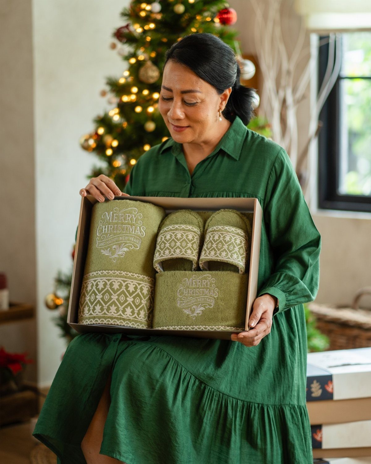 Woman holding a gift box of green Christmas themed tea towels near a tree.