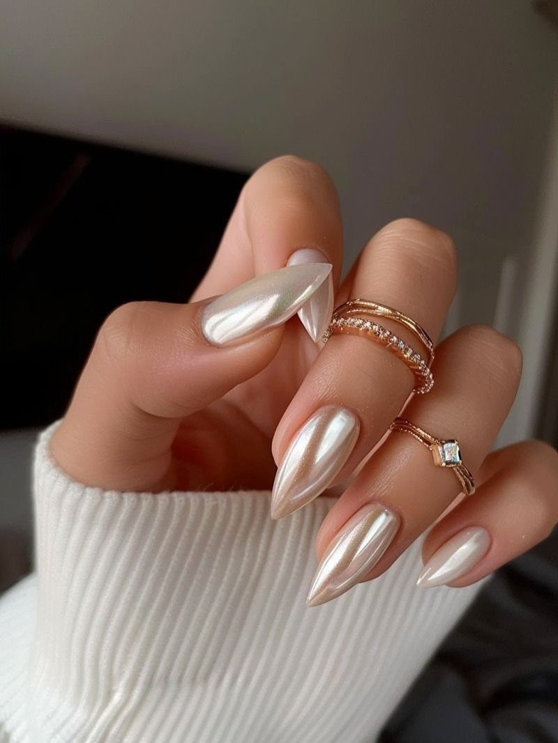 Almond-shaped nails with a pearlescent chrome finish and gold rings on fingers.