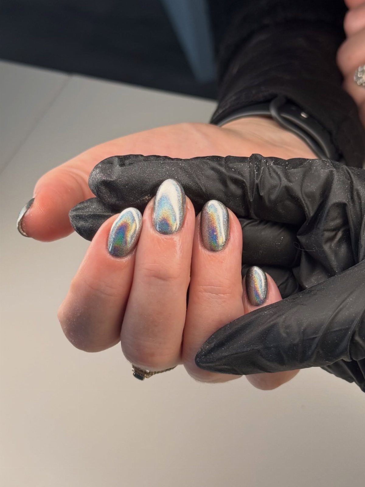 Holographic chrome nails with rainbow reflections held by a hand in black gloves.